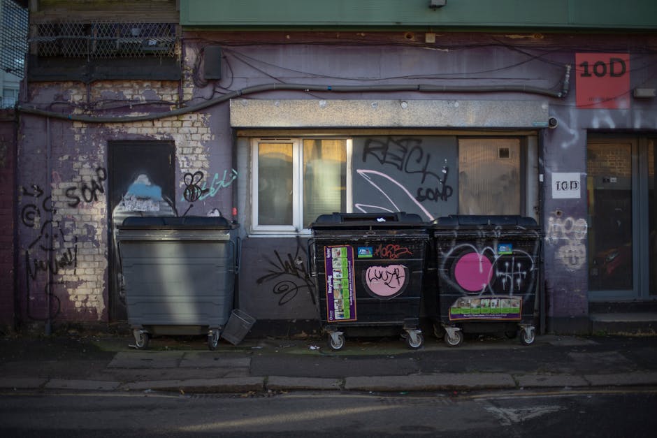 Three large wheelie bins are positioned along the sidewalk in front of a weathered building with a purple-painted brick wall. The first bin, on the left, is grey with a closed lid, and appears to be m