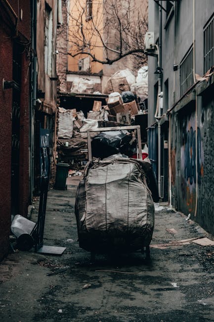 A scene of waste collection on a paved urban street at night, featuring three large, black plastic garbage bags filled with waste materials placed close to a black metal fence with vertical bars. The 