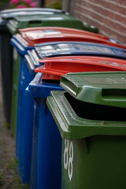 Five large black wheeled rubbish bins with yellow lids are positioned in a row against a plain, light gray wall, on a concrete surface. Each bin has a white oval label with printed identification numb
